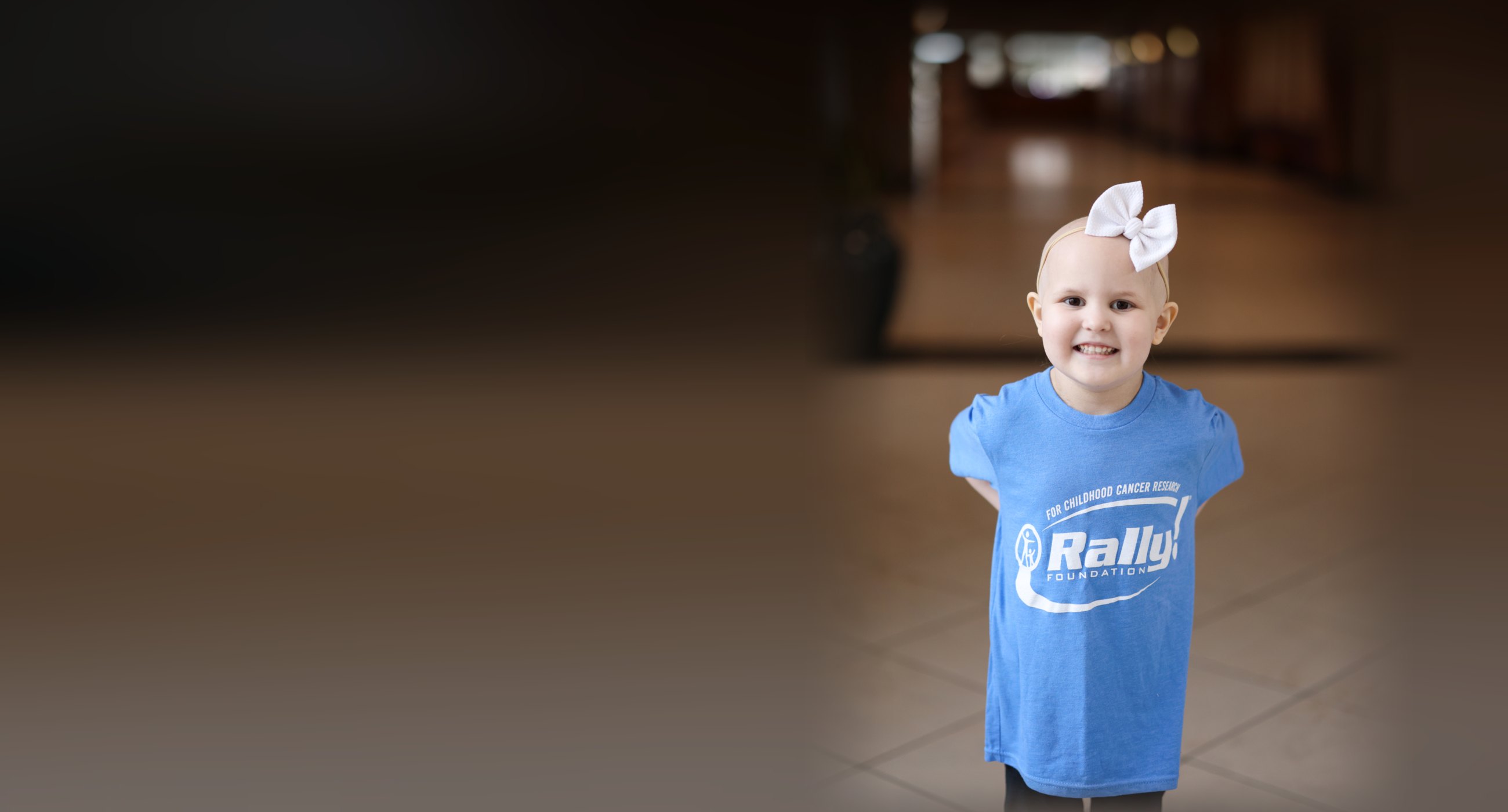 Nora, a young Rally Foundation patient, smiling in a hospital lobby