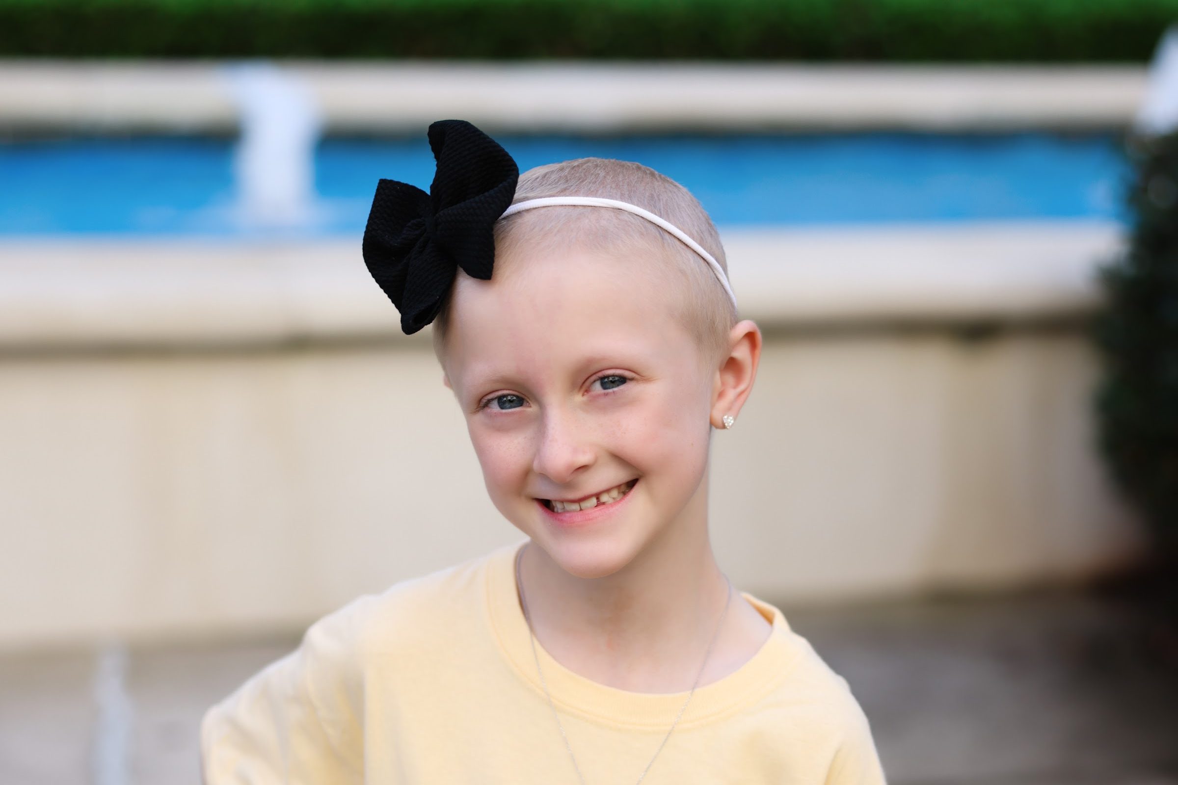 Natalee, a Rally Kid, smiling outdoors in a yellow Rally Foundation T-shirt and black bow headband